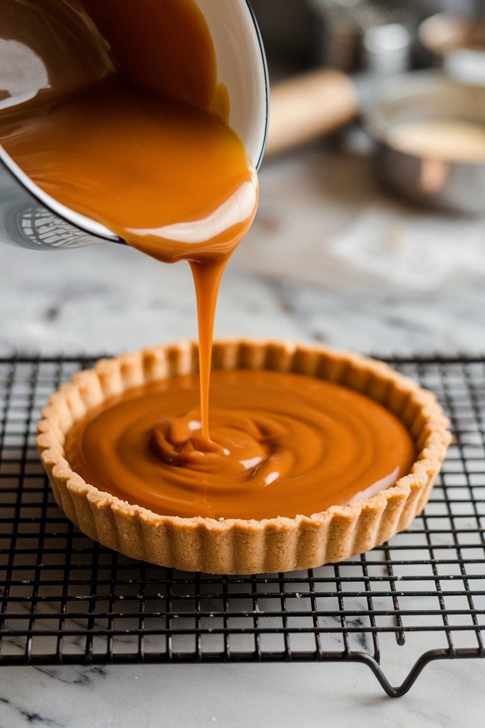 Rich caramel sauce being poured into a baked tart shell on a cooling rack, with the silky caramel spreading evenly.
