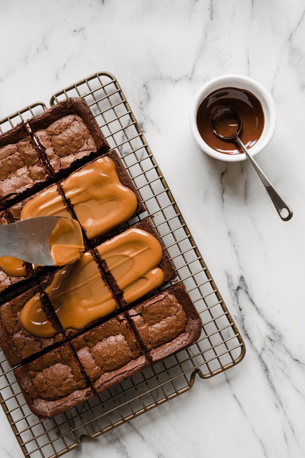 A cooling rack with sliced brownies partially coated in a thick layer of caramel being spread with a spatula. A small bowl of melted chocolate with a spoon is placed beside the rack on a marble surface.