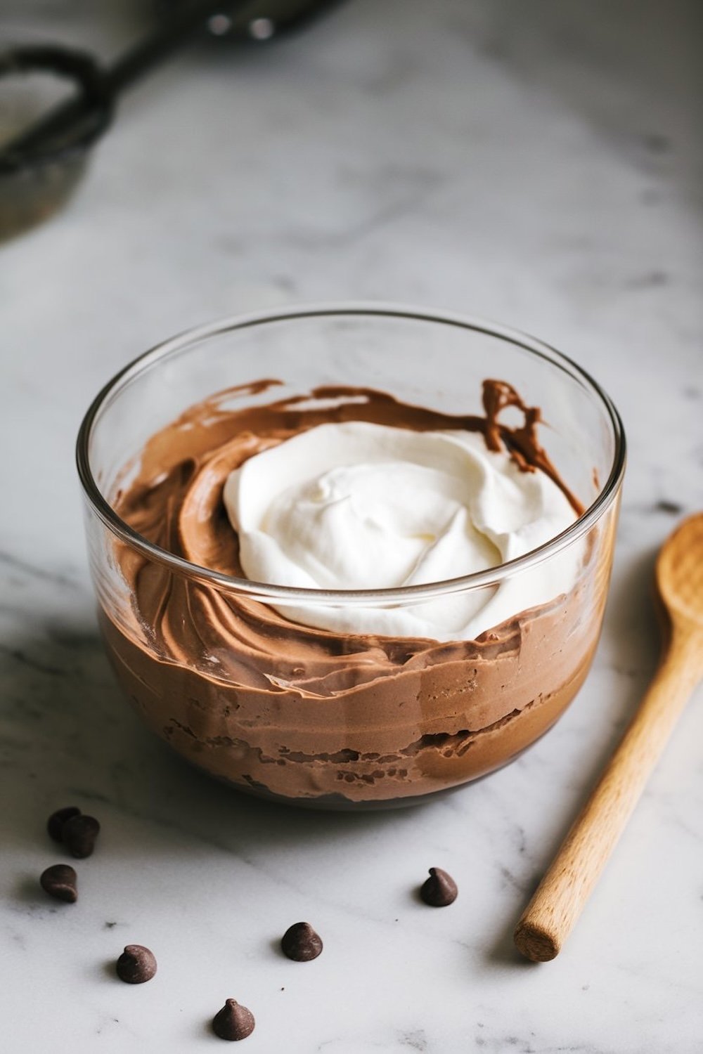A glass bowl filled with creamy chocolate mousse, topped with freshly whipped cream, surrounded by scattered chocolate chips on a marble surface, with a wooden spoon beside it.