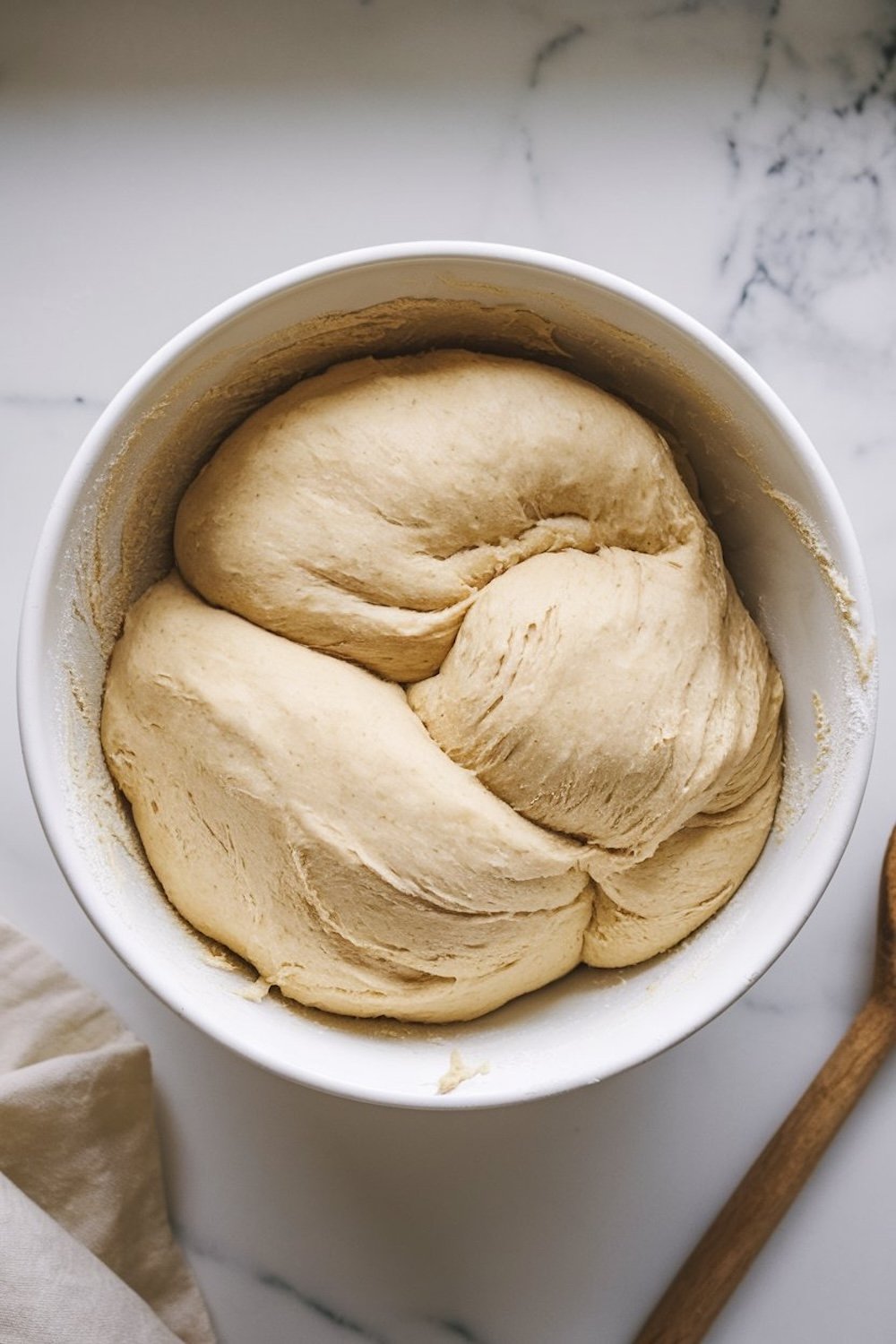 A bowl of proofed croissant dough with a soft, fluffy texture, resting on a marble countertop with a wooden spoon beside it.