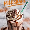 A creamy chocolate milkshake served in a tall glass, topped with whipped cream, chocolate shavings, and a drizzle of chocolate syrup. A green and white striped straw is placed in the milkshake. The background features cocoa powder and chocolate syrup splatters.