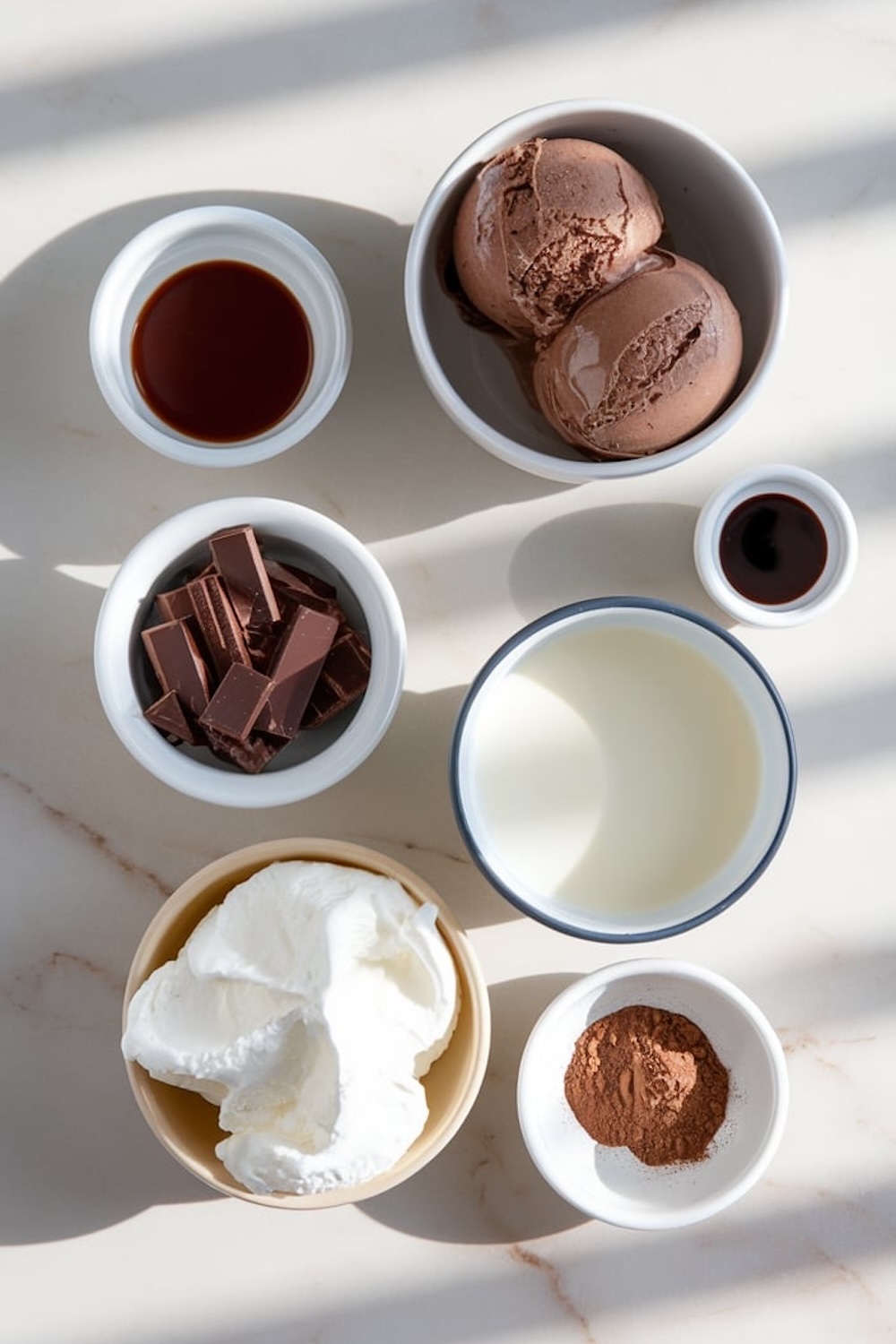 A flat-lay of ingredients for making a chocolate milkshake, including scoops of chocolate ice cream, chopped chocolate, milk, cocoa powder, whipped cream, vanilla extract, and chocolate syrup, arranged in small bowls on a marble surface.