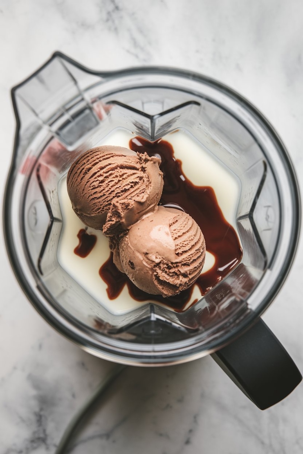 A blender jar with two scoops of chocolate ice cream, milk, and chocolate syrup, ready for blending. The glass jar sits on a marble countertop.