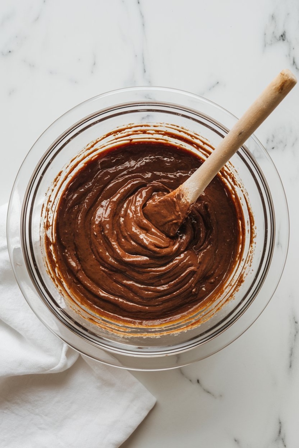 A glass bowl filled with glossy chocolate brownie batter being mixed with a wooden spatula, placed on a marble countertop.
