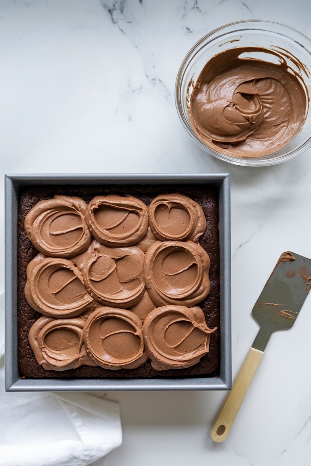 A baking pan with freshly spread chocolate mousse over a brownie base. A bowl of mousse and a spatula sit beside the pan on a marble countertop.