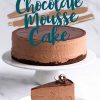 A full chocolate mousse cake with a glossy chocolate ganache top displayed on a cake stand. A slice is served on a plate below, showcasing the layers of chocolate crust, airy mousse, and smooth ganache. Decorative chocolate curls garnish the slice, adding a touch of elegance to the rich dessert presentation. Text overlay highlights “Chocolate Mousse Cake.”