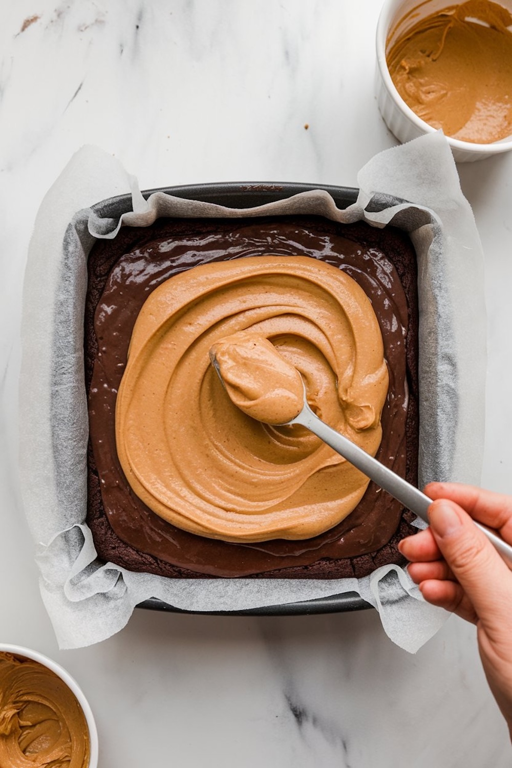A baking pan lined with parchment paper, filled with chocolate batter topped with a creamy peanut butter layer being spread with a spoon. A bowl of peanut butter is visible nearby, highlighting the layering process for brownies.