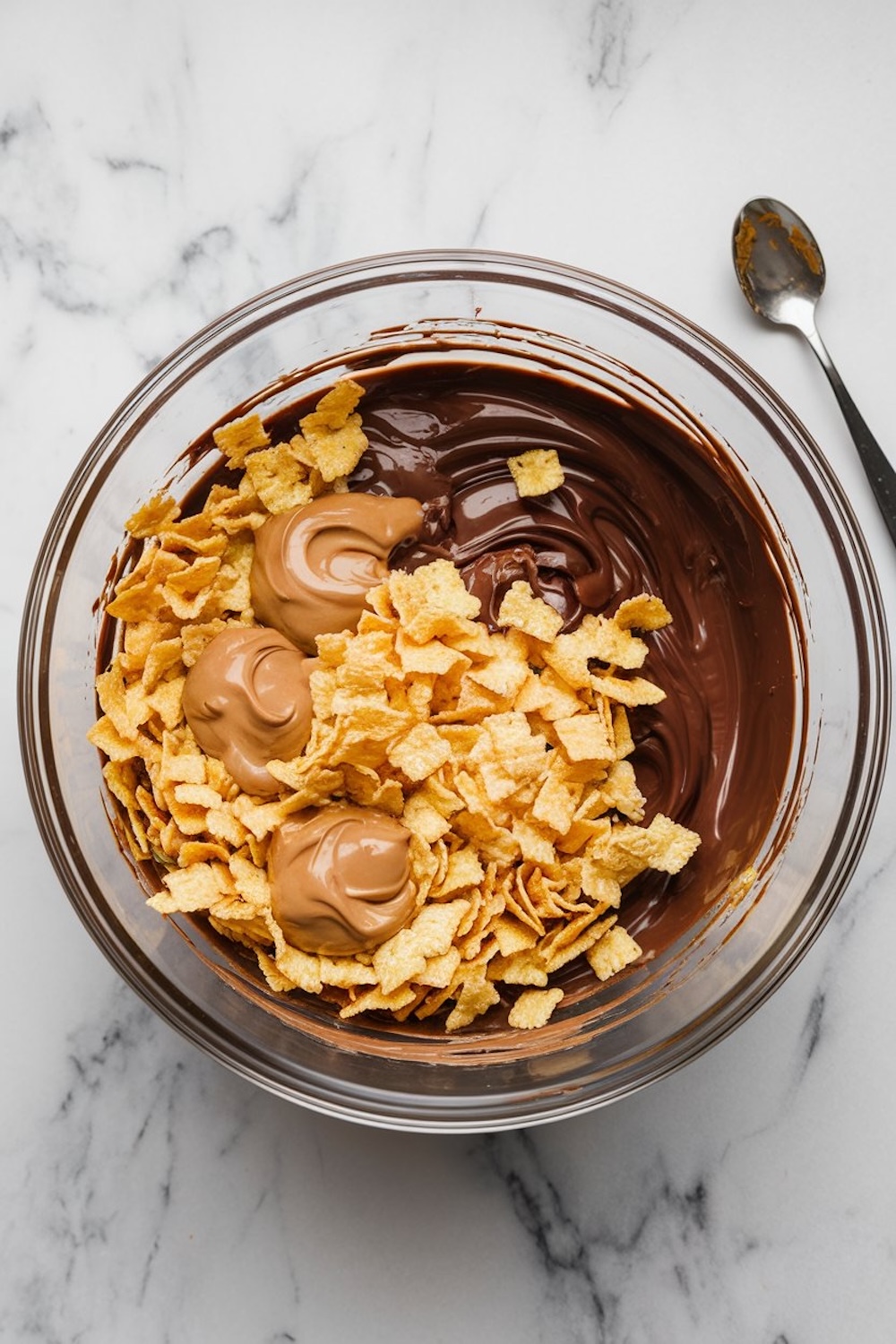Bowl of melted chocolate and peanut butter combined with crispy cereal squares, featuring dollops of additional peanut butter for mixing, all set on a marble surface with a spoon nearby.