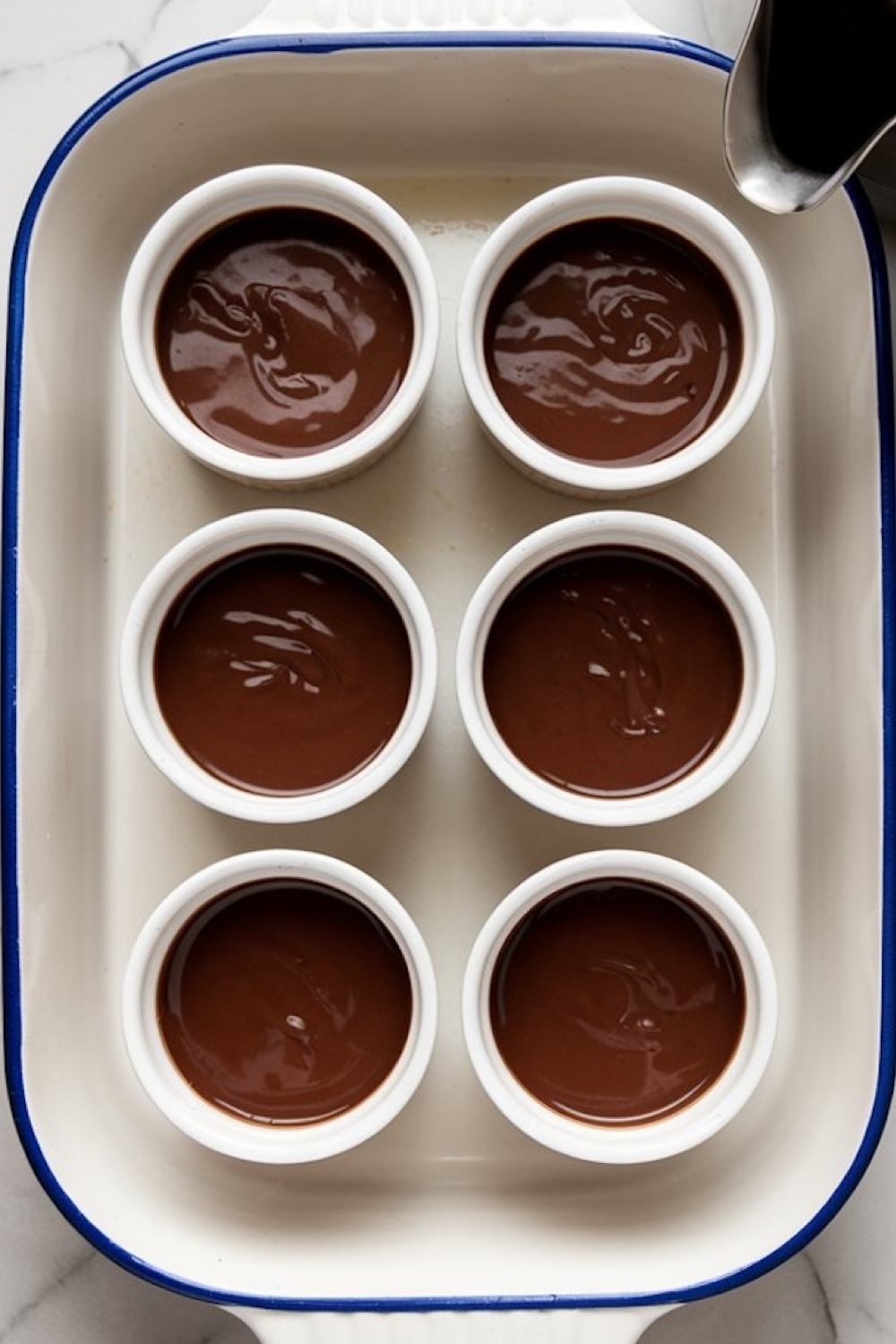Six white ramekins filled with smooth chocolate pot de crème placed inside a white ceramic baking dish with blue edges, resting on a marble surface.