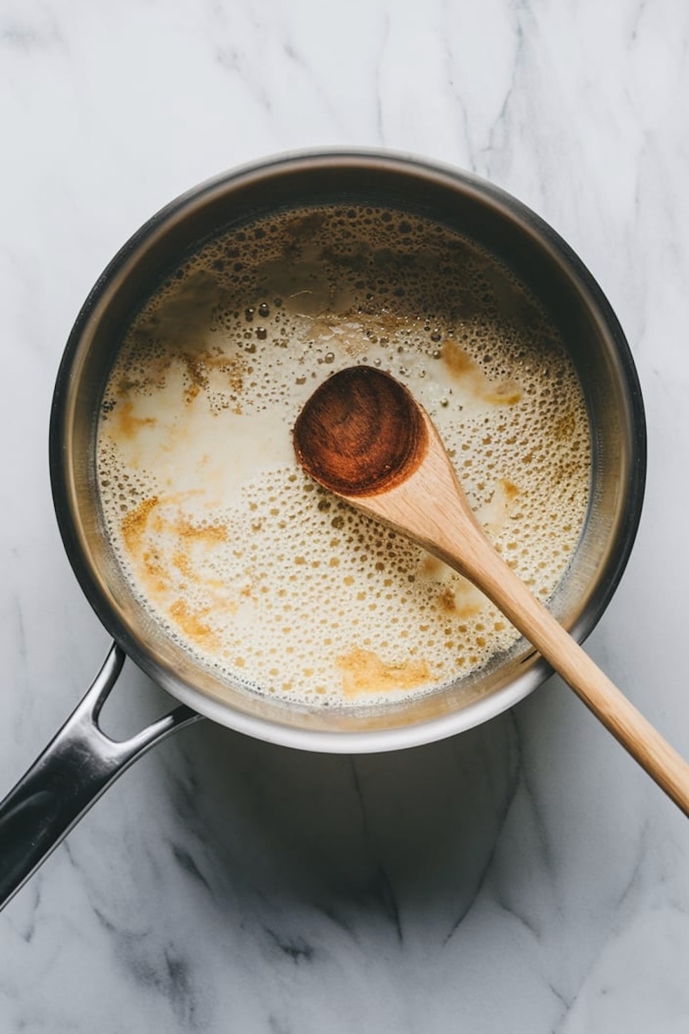 A saucepan filled with bubbling milk and vanilla mixture, being stirred with a wooden spoon. The pan rests on a marble surface, showing the pudding preparation in progress.