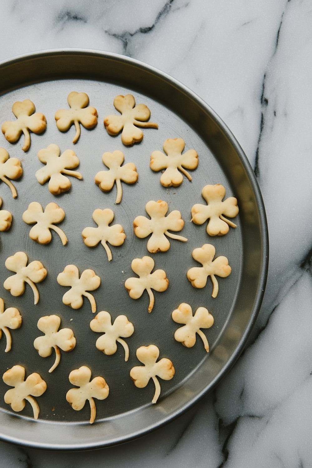 A baking tray filled with baked shamrock-shaped cookie toppers, placed on a white marble surface. The golden-brown edges of the shamrocks showcase their crisp texture.