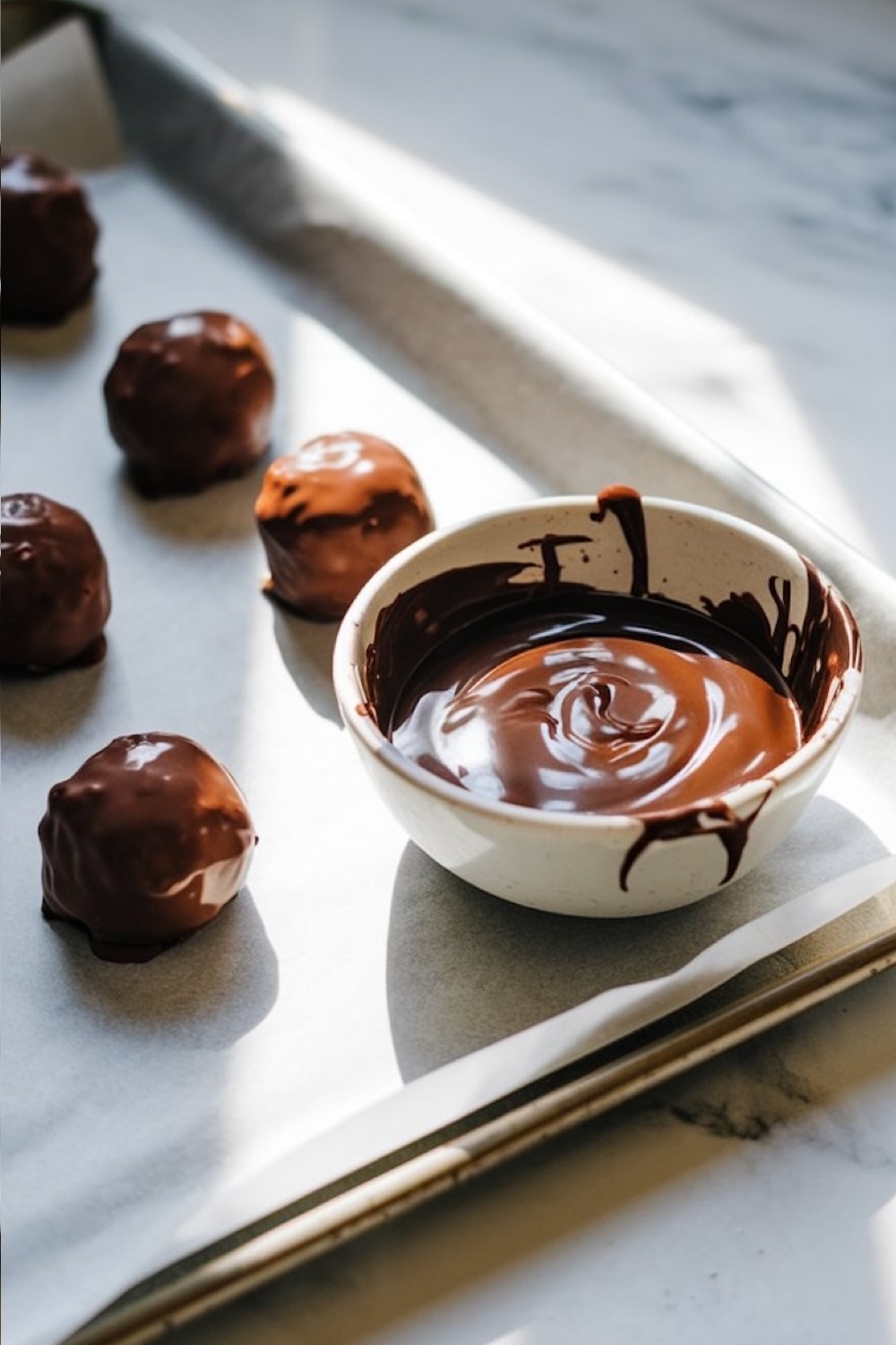 Freshly coated chocolate truffles cooling on a parchment-lined baking sheet in soft natural light. A small bowl of melted chocolate sits nearby, with remnants of chocolate dripping down the sides, emphasizing the process of dipping the truffles to achieve their glossy finish.