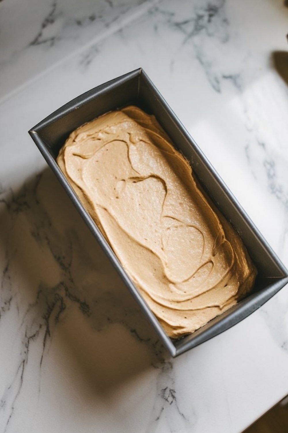 A loaf pan filled with creamy banana bread batter, ready to be baked. The pan is placed on a marble countertop, lit with soft natural light.