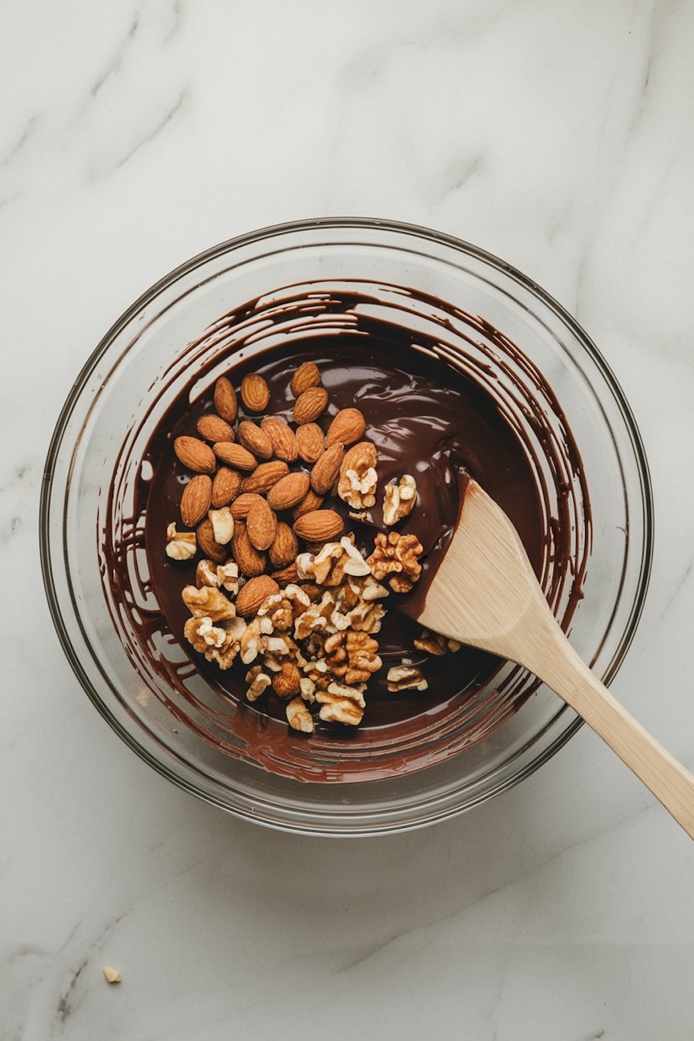 A glass bowl of melted dark chocolate with whole almonds and walnut halves added on top. A wooden spatula lies inside, ready for mixing. The setup rests on a white marble surface.