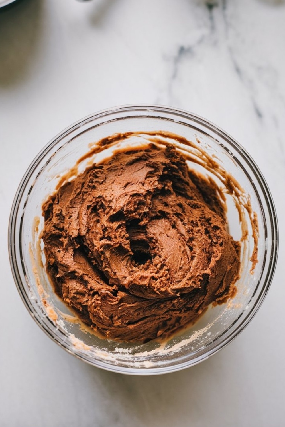 A glass bowl filled with dark chocolate cookie dough. The dough has a creamy, thick texture with a rich brown color, sitting on a marble surface, ready for baking.