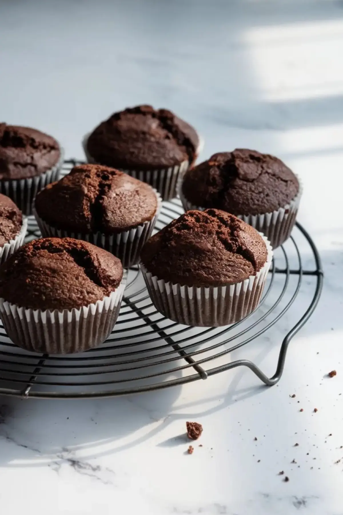 Freshly baked chocolate cupcakes on a black wire cooling rack, with cracked tops and a rich, dark color. Sunlight casts shadows on a white marble countertop, with a few crumbs scattered nearby.