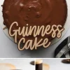 A rich Guinness chocolate cake covered in glossy chocolate ganache sits on a white plate. The text “Guinness Cake” overlays the image in elegant script. Below, two slices of the layered cake reveal moist chocolate sponge with creamy chocolate filling, drizzled with ganache. A cup of dark stout beer is in the background.