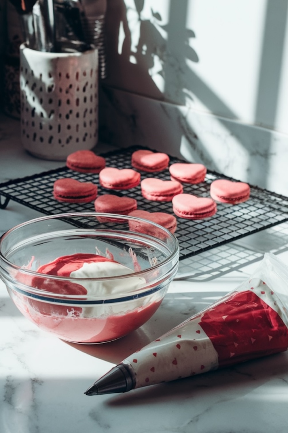 A baking setup in natural light showcasing heart-shaped macarons cooling on a black wire rack. In the foreground, a clear bowl contains red and white macaron filling, and a decorated piping bag with red heart patterns rests beside it on a marble countertop.
