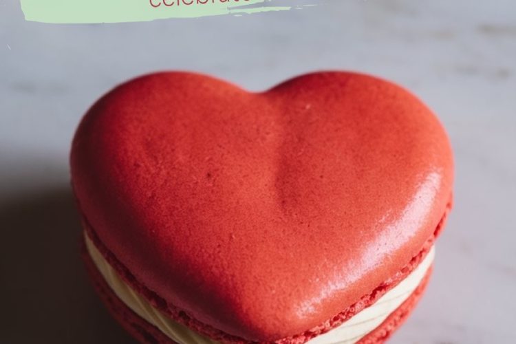 A single vibrant red heart-shaped macaron with white filling is centered on a white marble surface, next to the edge of a white plate with gold trim. Overlaid text reads “Heart-shaped Macarons - Celebrate Love Simply.”