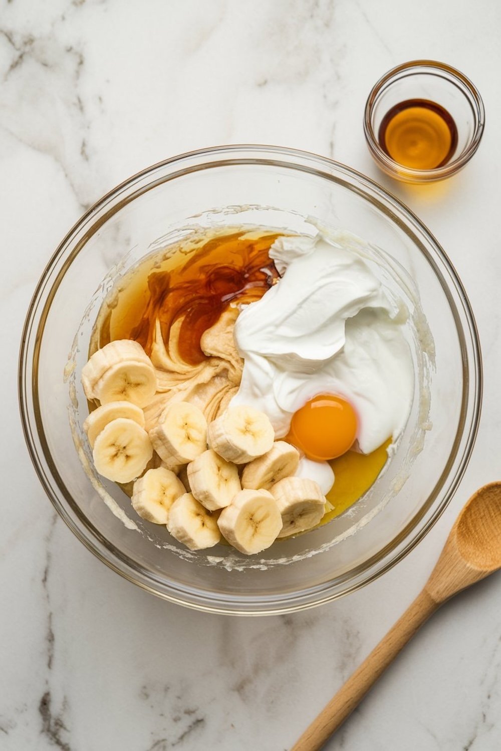 A glass mixing bowl on a white marble surface filled with banana bread ingredients, including sliced bananas, an egg, yogurt, peanut butter, and vanilla extract. A small bowl of vanilla and a wooden spoon are placed nearby.