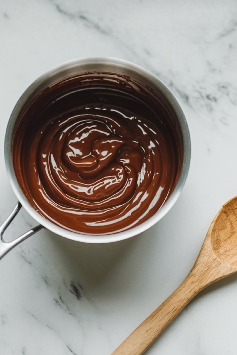 Smooth and glossy melted chocolate in a saucepan on a marble countertop, with a wooden spoon partially visible.