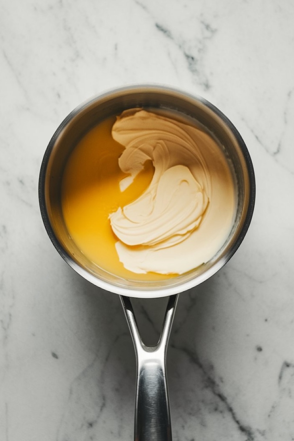 Saucepan containing melted butter and cream, creating a smooth, pale yellow mixture on a marble surface.