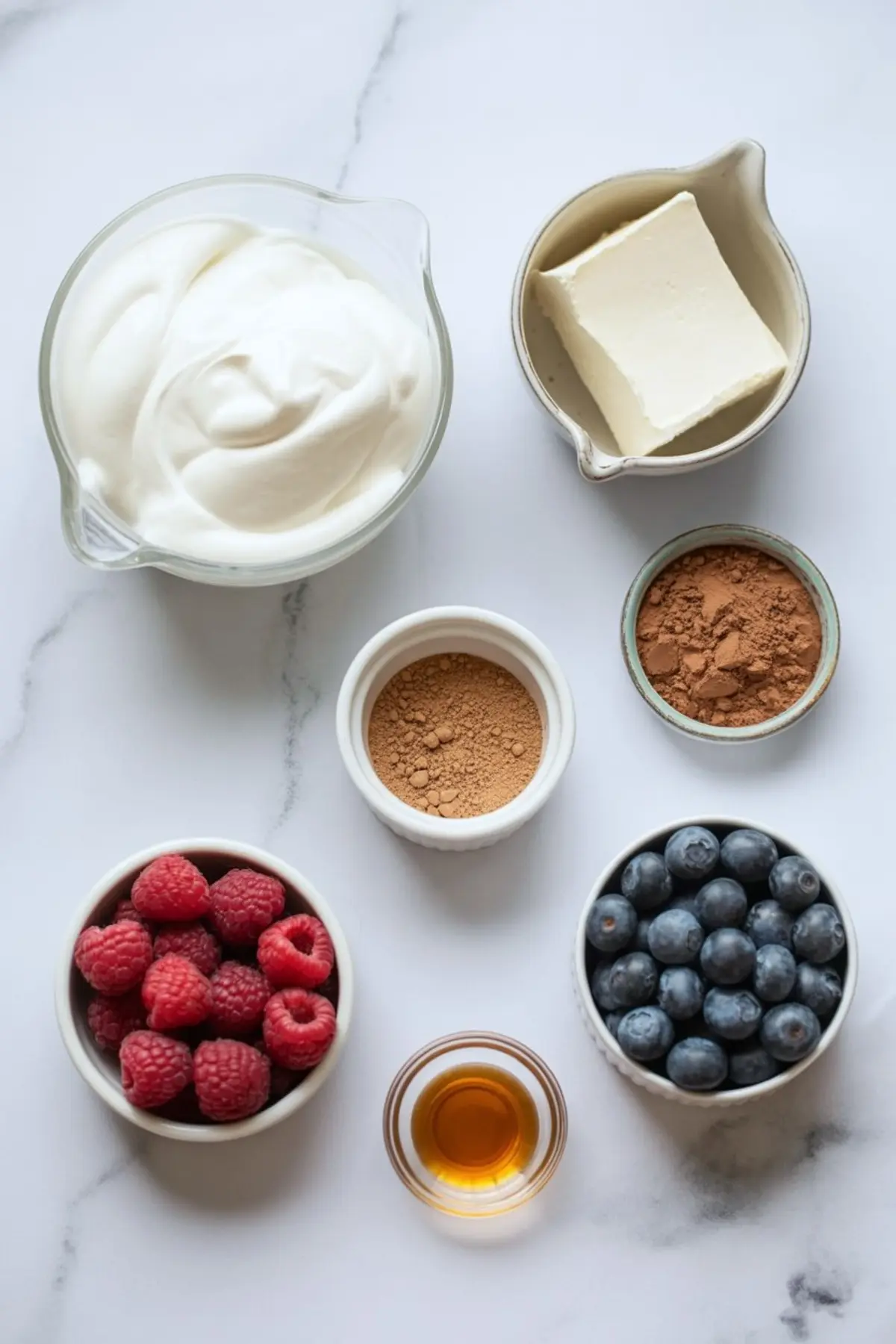 Flat lay of chocolate mousse ingredients on a white marble background, including whipped cream, cream cheese, cocoa powder, cacao powder, fresh raspberries, blueberries, vanilla extract, and a bowl of Greek yogurt.
