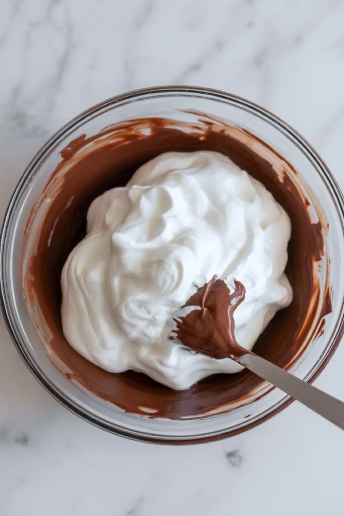 Chocolate mousse mixture in progress with fluffy whipped cream being folded into melted chocolate in a clear glass bowl.
