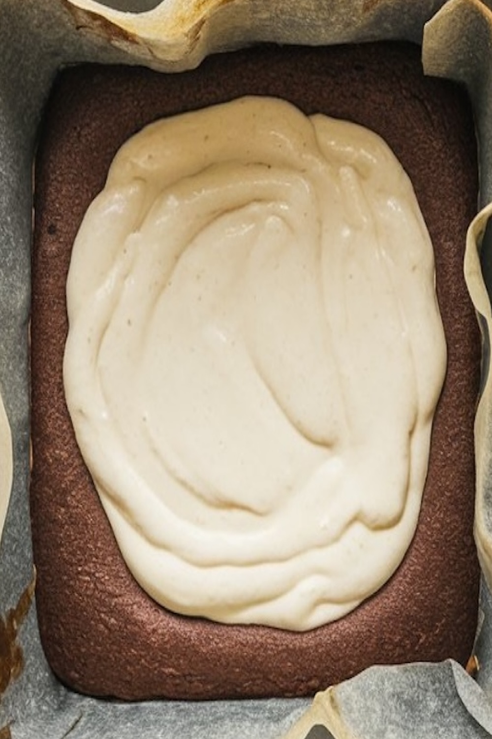 A baking pan lined with parchment paper, filled with chocolate brownie batter topped with a layer of creamy cheesecake filling, ready for swirling. The surface appears smooth and prepped for baking.