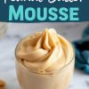 A glass of creamy peanut butter mousse with a swirled, fluffy texture. The dessert sits on a marble surface with text overlay reading “Peanut Butter Mousse” in bold, colorful font. A blue napkin and peanuts are visible in the background.