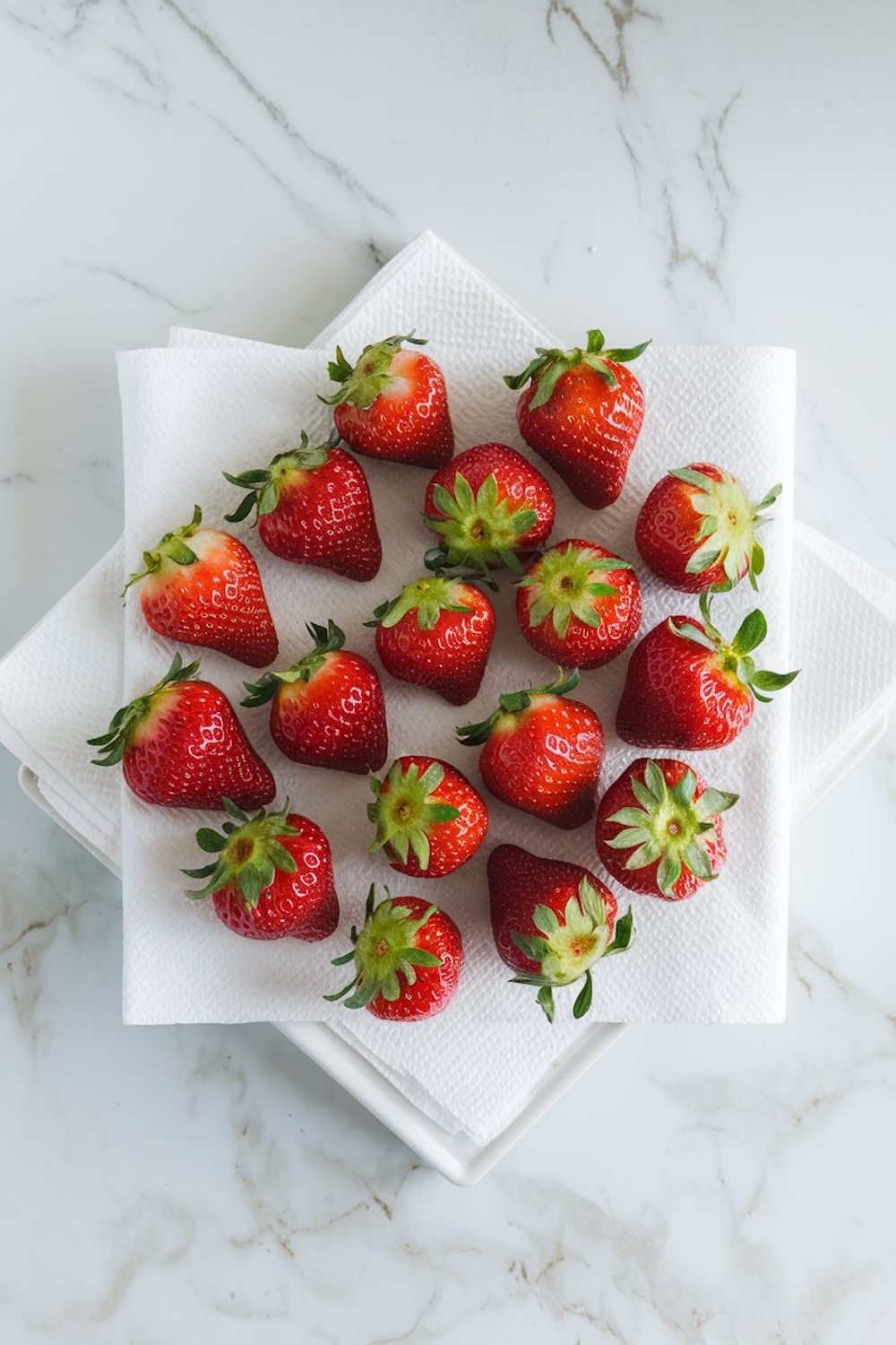 Freshly washed strawberries drying on a paper towel, arranged neatly on a white plate with a marble background.