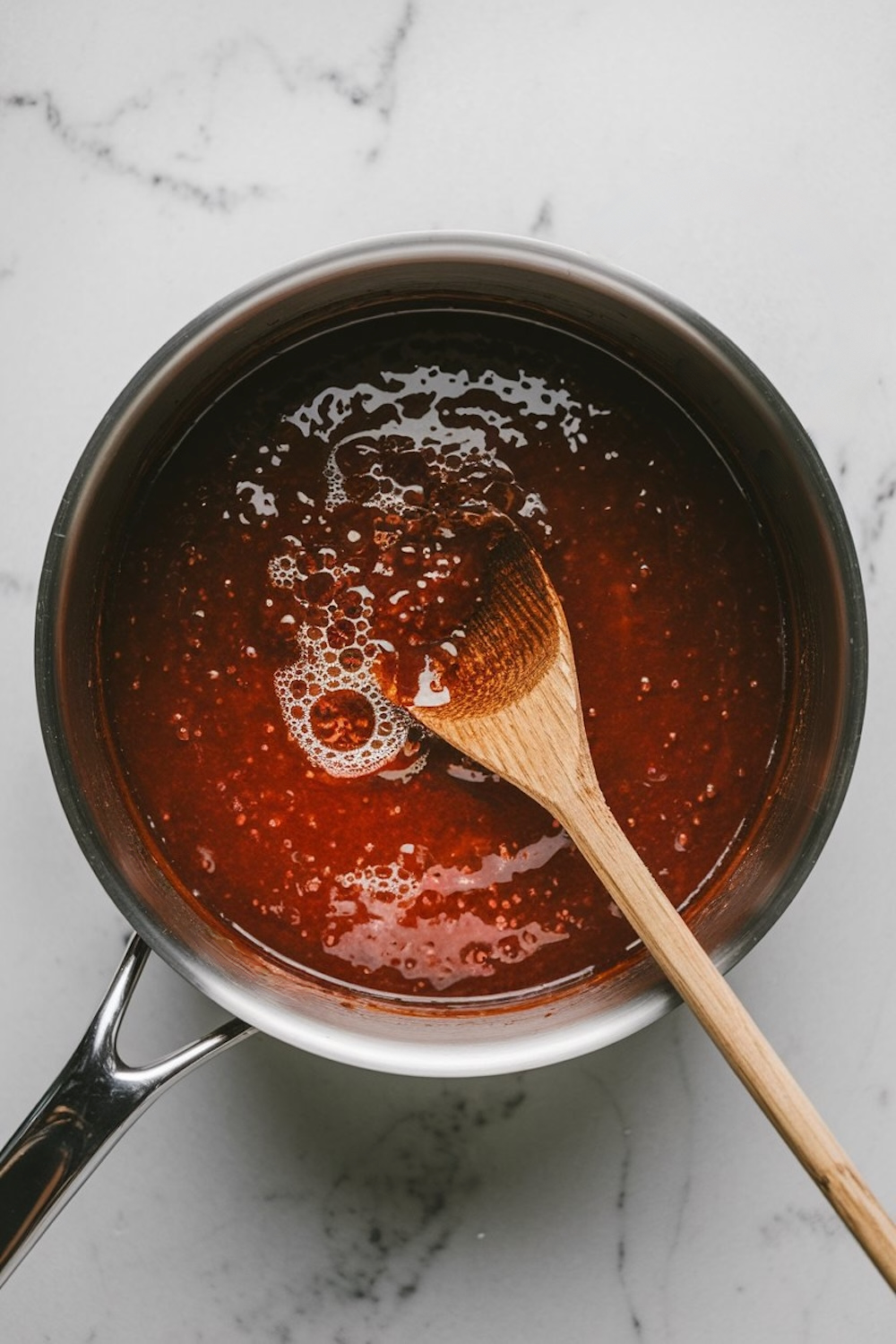 Overhead view of a saucepan filled with bubbling sorrel jam in the cooking process, showcasing its rich, deep red color and thick texture. A wooden spoon rests in the mixture, indicating the jam is being stirred to achieve the perfect consistency. The saucepan is placed on a smooth white marble surface, emphasizing the vibrant hues of the jam.