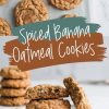A plate of spiced banana oatmeal cookies stacked with one broken cookie leaning against the stack, showing the soft and moist interior. The background includes blurred stacks of cookies for added depth.