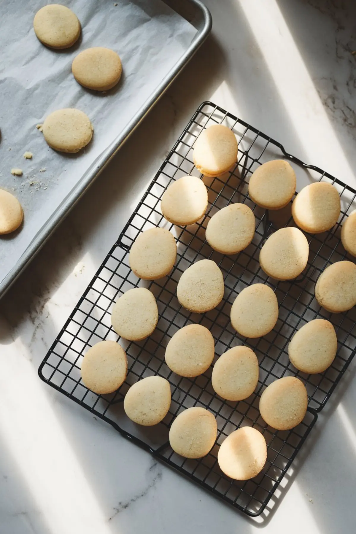 Freshly baked, golden egg-shaped sugar cookies cooling on a black wire rack and a parchment-lined baking sheet. Soft natural light highlights their texture.