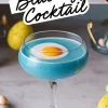 Close-up of a blue egg cocktail in a coupe glass with a white layer and an apricot garnish resembling a fried egg. The elegant glass is surrounded by pastel Easter eggs and festive decorations, emphasizing a fun and creative Easter-inspired drink presentation.
