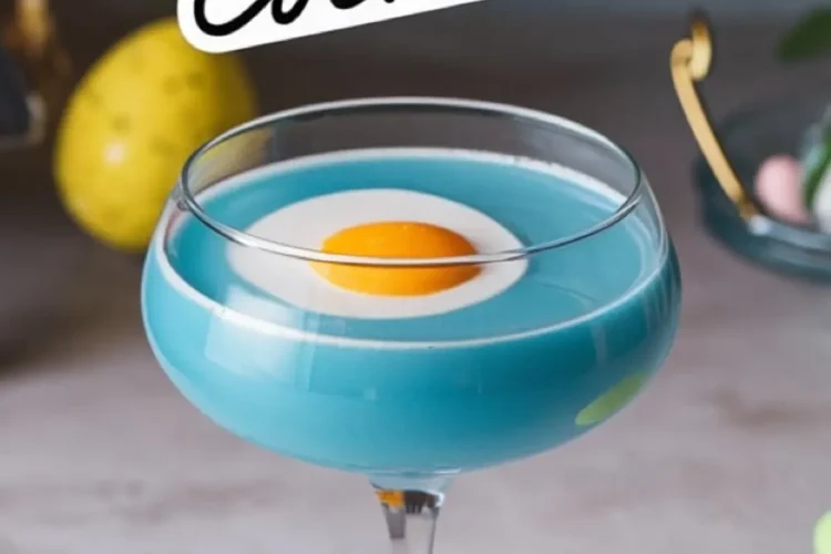 Close-up of a blue egg cocktail in a coupe glass with a white layer and an apricot garnish resembling a fried egg. The elegant glass is surrounded by pastel Easter eggs and festive decorations, emphasizing a fun and creative Easter-inspired drink presentation.