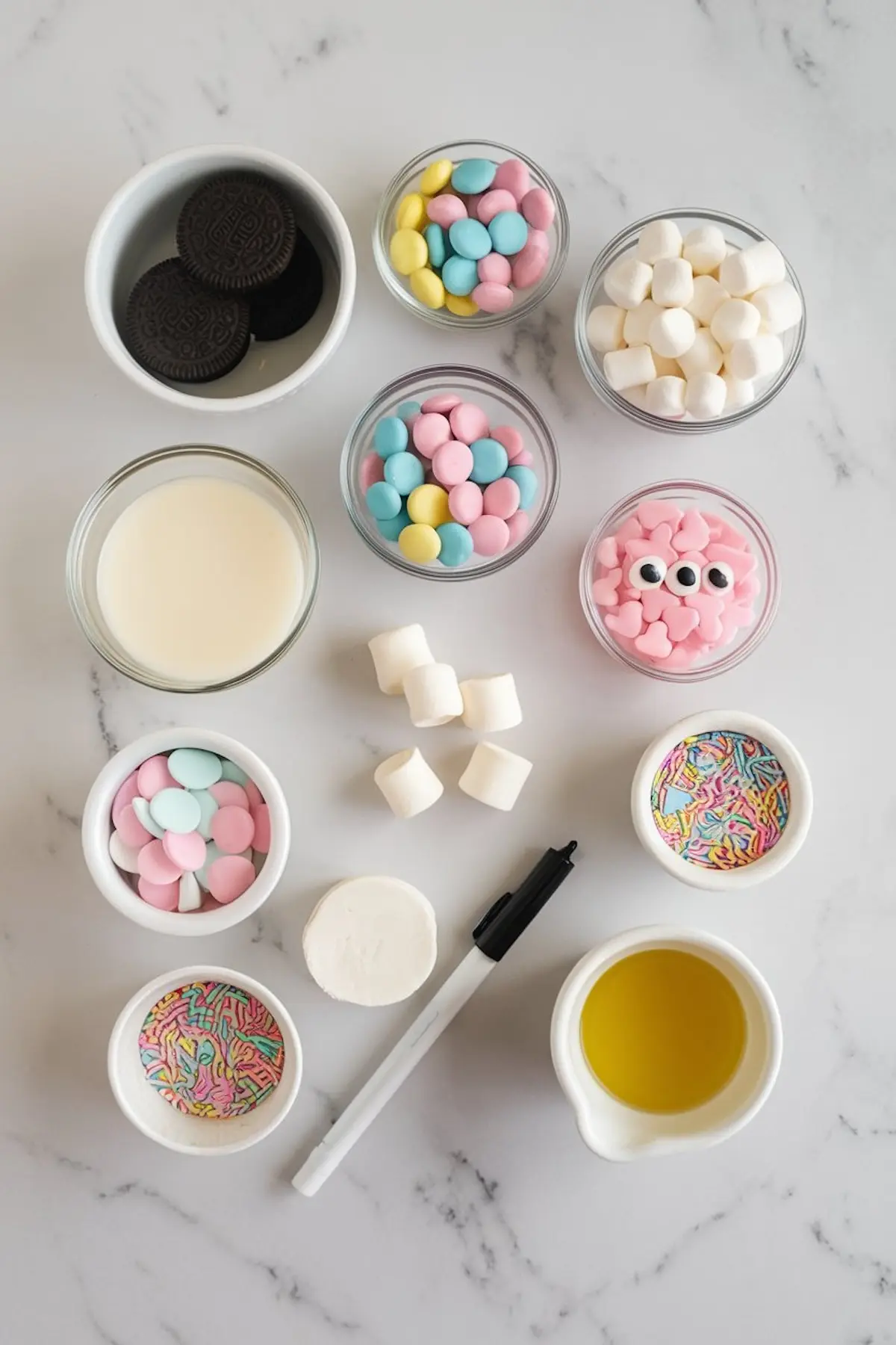 Flat lay of ingredients on a white marble surface for making decorated Oreo pops. Items include chocolate sandwich cookies, pastel candy melts, mini marshmallows, candy eyes, heart-shaped sprinkles, rainbow sprinkles, heavy cream, oil, a black edible marker, and lollipop sticks.