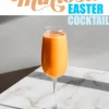 Carrot mimosa in a champagne flute on a marble surface, illuminated by natural light. The vibrant orange drink contrasts with the sleek, elegant background. Bold text above reads “Carrot Mimosa Easter Cocktail,” emphasizing its festive, springtime appeal.