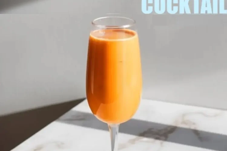 Carrot mimosa in a champagne flute on a marble surface, illuminated by natural light. The vibrant orange drink contrasts with the sleek, elegant background. Bold text above reads “Carrot Mimosa Easter Cocktail,” emphasizing its festive, springtime appeal.