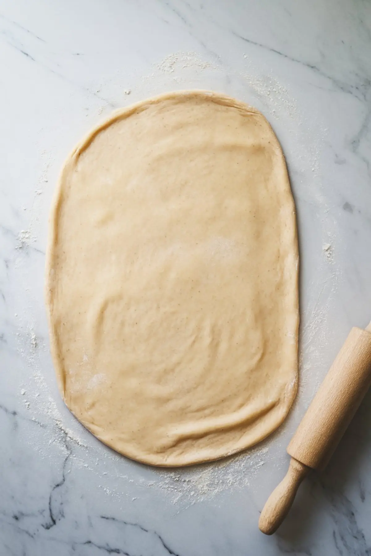 Rolled-out dough on a floured marble surface, with a wooden rolling pin resting beside it. The dough is evenly flattened, ready for the filling to be spread before shaping into cinnamon rolls.