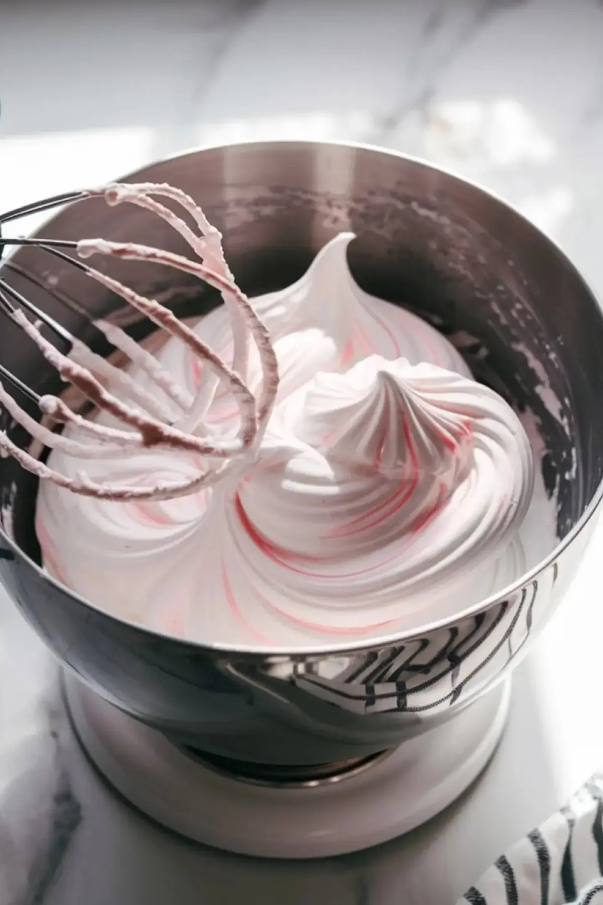 A stainless steel mixing bowl filled with freshly whipped raspberry meringue, featuring soft pink swirls. A whisk attachment rests above the bowl, coated in the glossy mixture. The marble countertop underneath provides a clean, bright background.