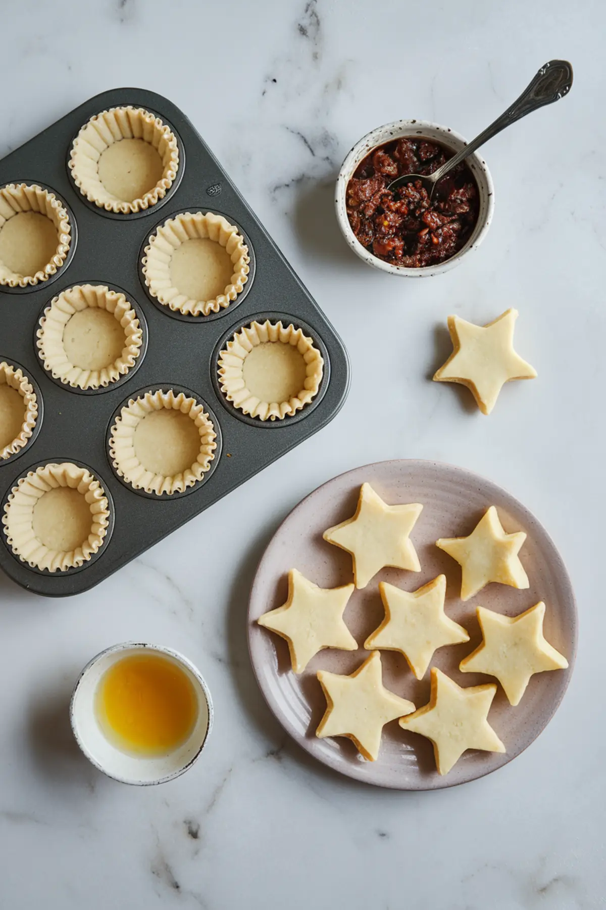 A baking scene with a muffin tin lined with unbaked pastry shells, a plate of star-shaped pastry cutouts, a bowl of dark mincemeat filling with a spoon, and a small dish of egg wash, all arranged on a white marble surface.