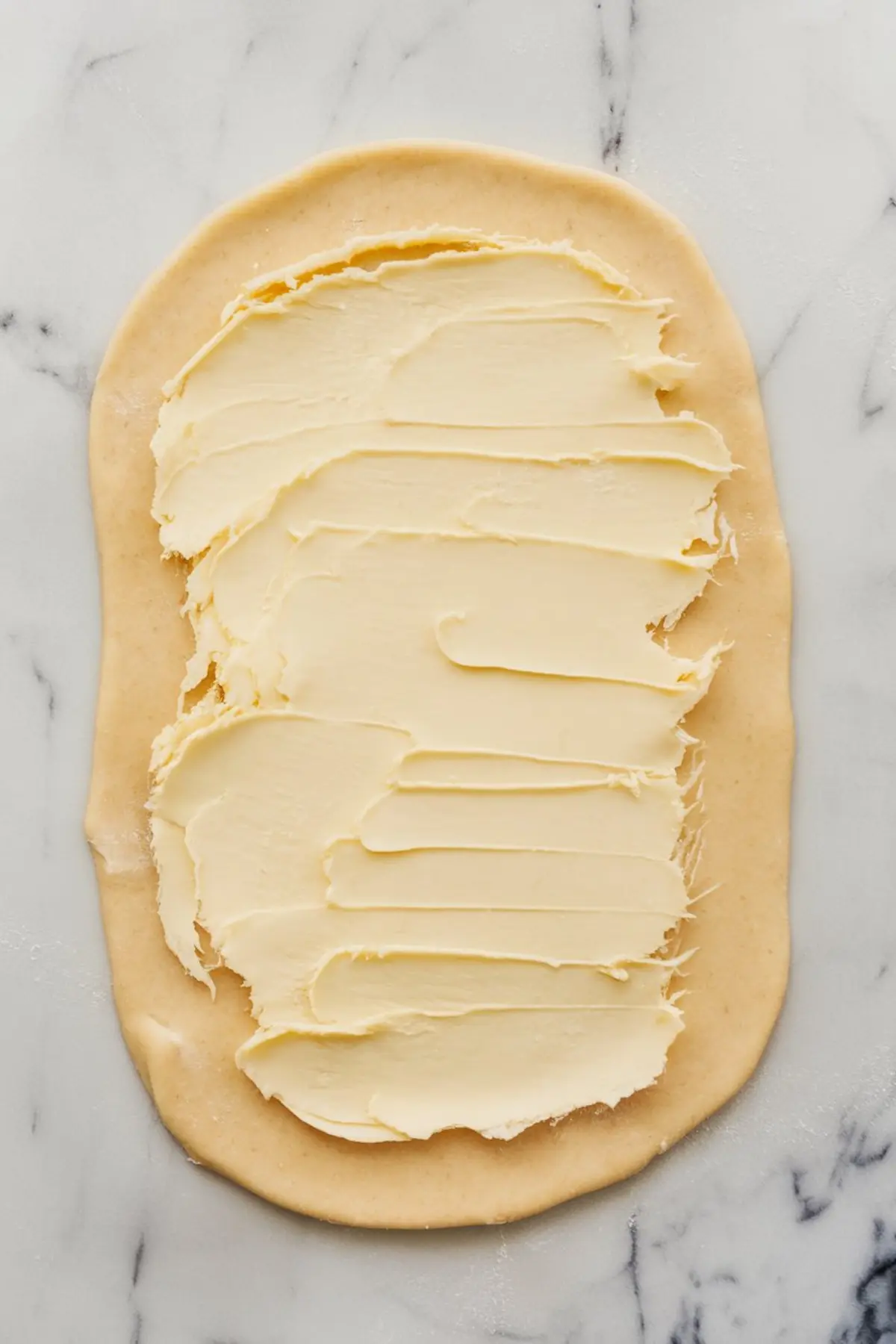 A sheet of dough rolled out on a marble surface with a thick layer of softened butter spread evenly across the top. The dough has slightly uneven edges, and the butter is creamy and smooth, ready for the next step in the baking process.