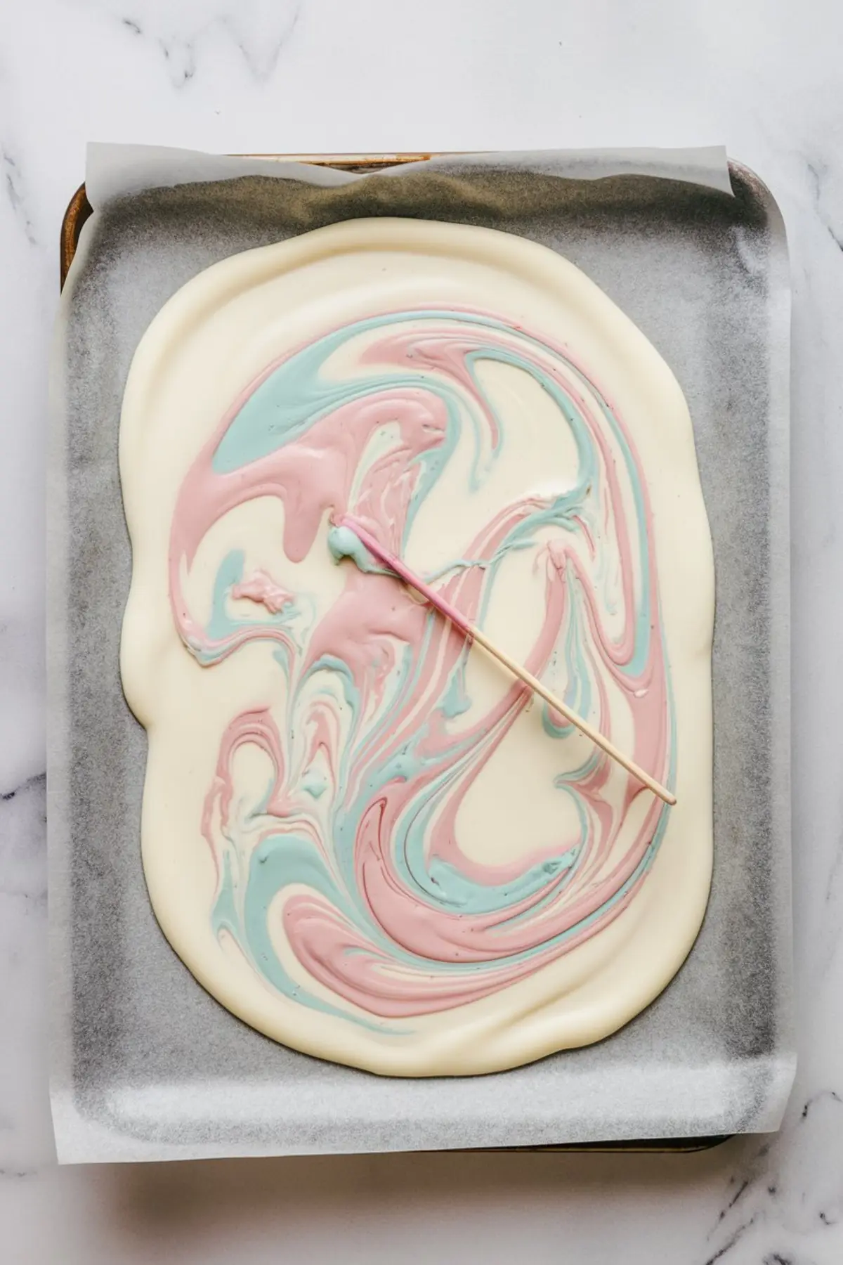 A baking sheet lined with parchment paper holding a base of melted white chocolate with pastel pink and blue swirls. A wooden skewer is being used to create a marbled pattern in the chocolate.