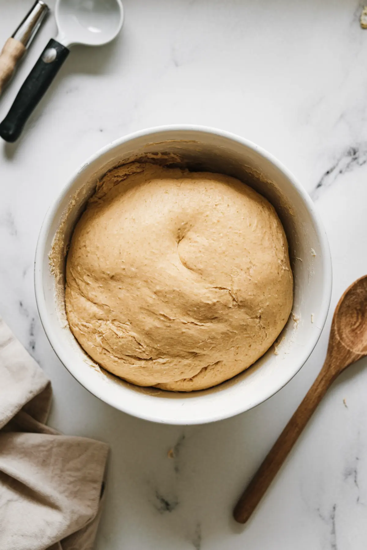 A bowl of risen cinnamon roll dough with a smooth, elastic surface, sitting on a white marble countertop. A wooden spoon and measuring tools are in the background, suggesting the next step in the baking process.
