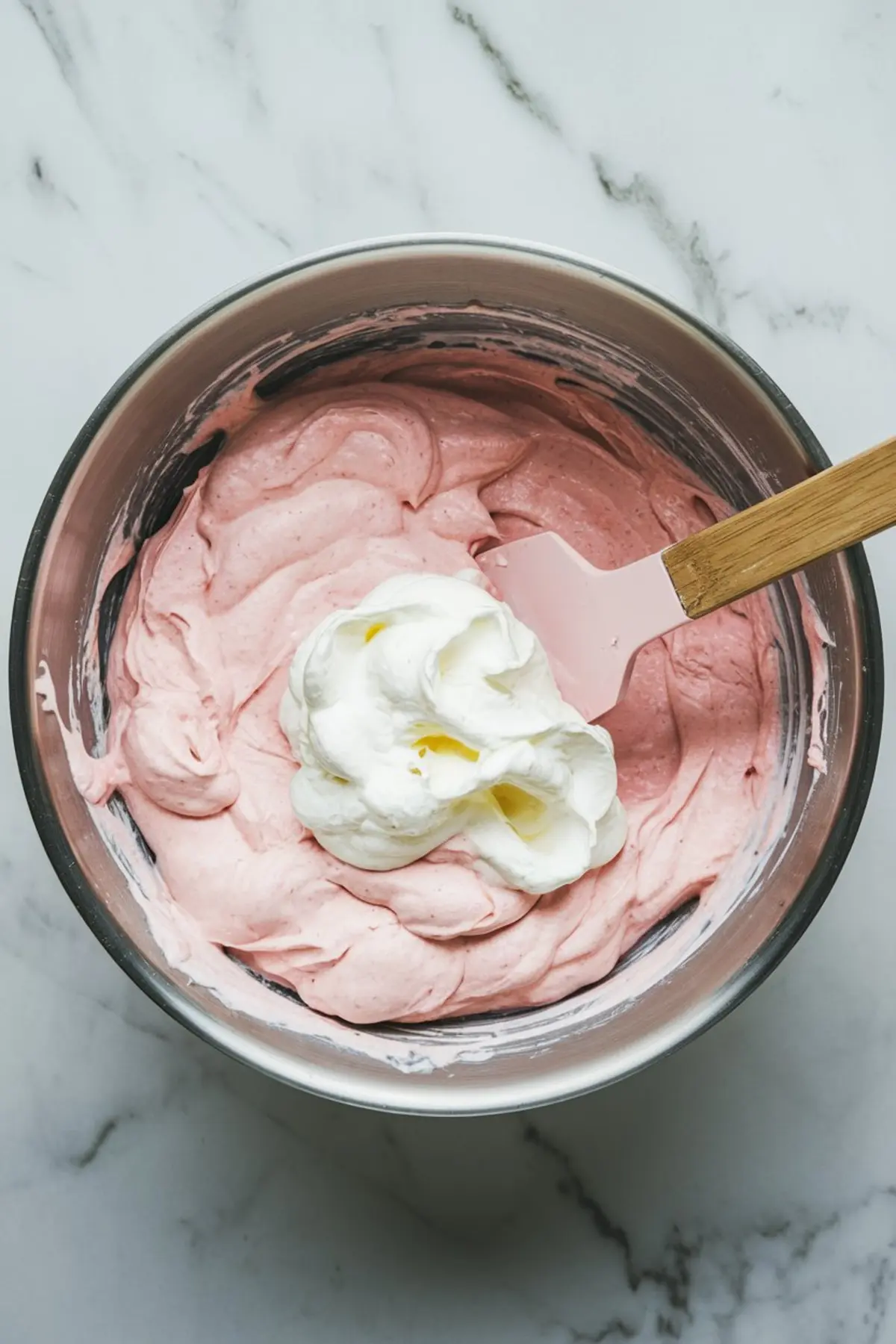 A metal mixing bowl filled with pink Easter dip, with a dollop of whipped topping being folded in using a spatula. The bowl rests on a white marble countertop.