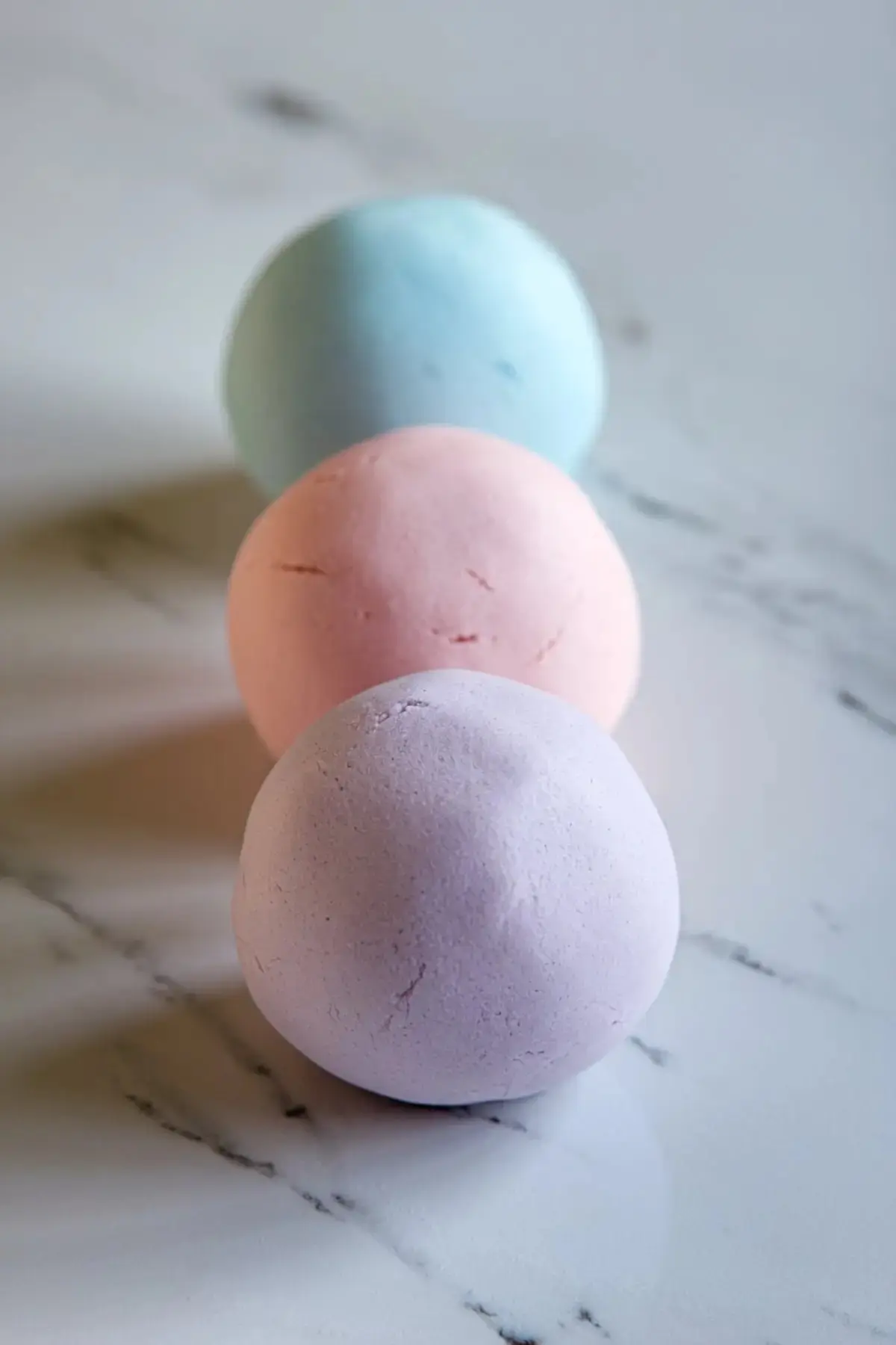 Three balls of pastel-colored fondant or cookie dough in soft lavender, blush pink, and baby blue, neatly lined up on a marble surface. The texture is smooth, and the colors are vibrant.