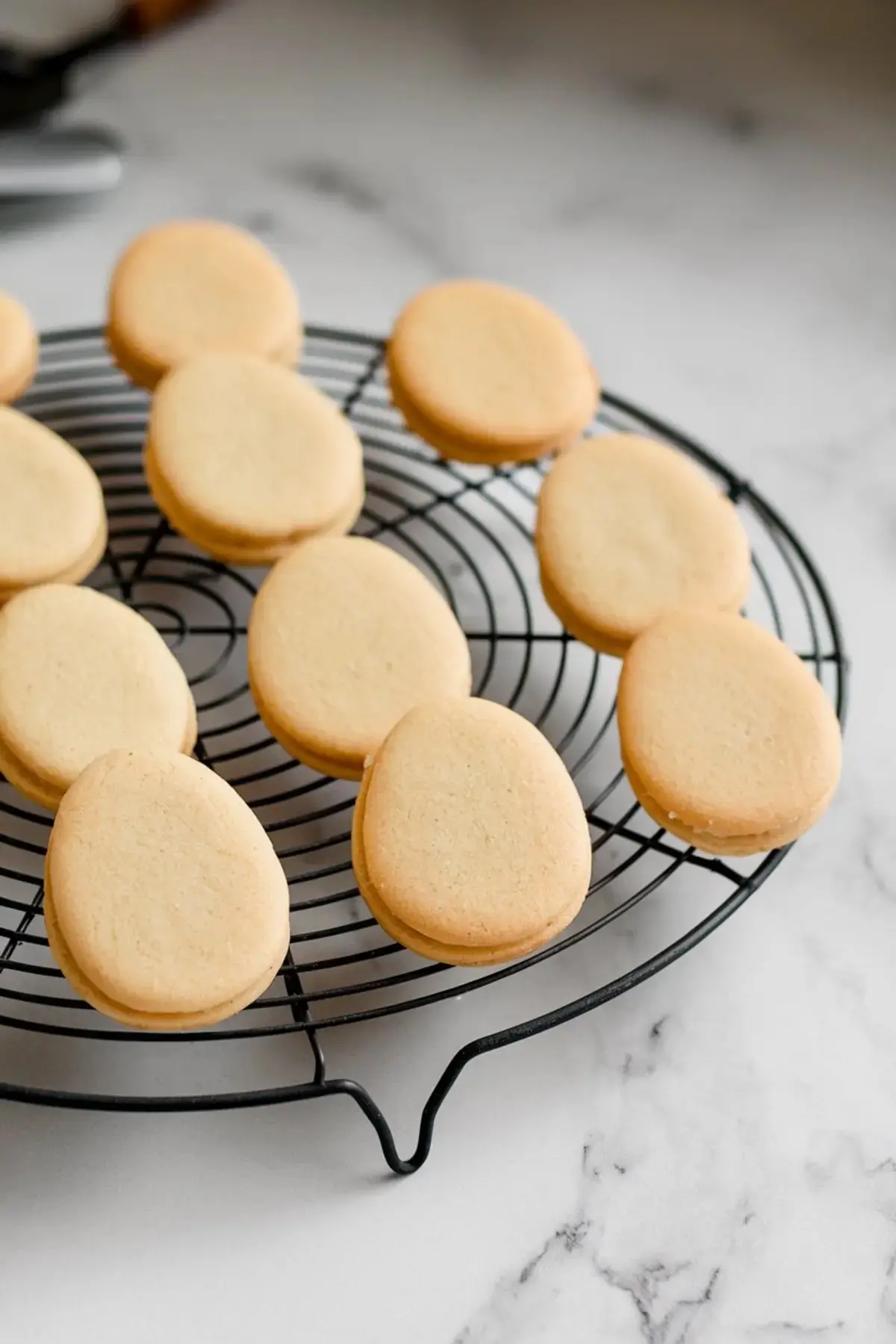 A batch of freshly baked golden brown egg-shaped sugar cookies cooling on a black wire rack. The cookies have a uniform shape and a smooth surface, ideal for decorating.