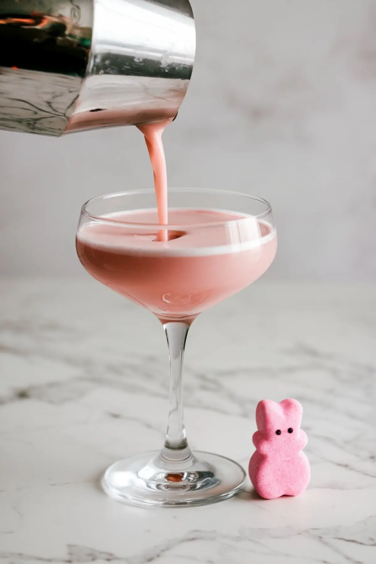 Pink Easter Peep martini being poured into a coupe glass from a silver cocktail shaker. A pink bunny-shaped marshmallow sits beside the glass, complementing the creamy pastel drink, ideal for an Easter-themed party.