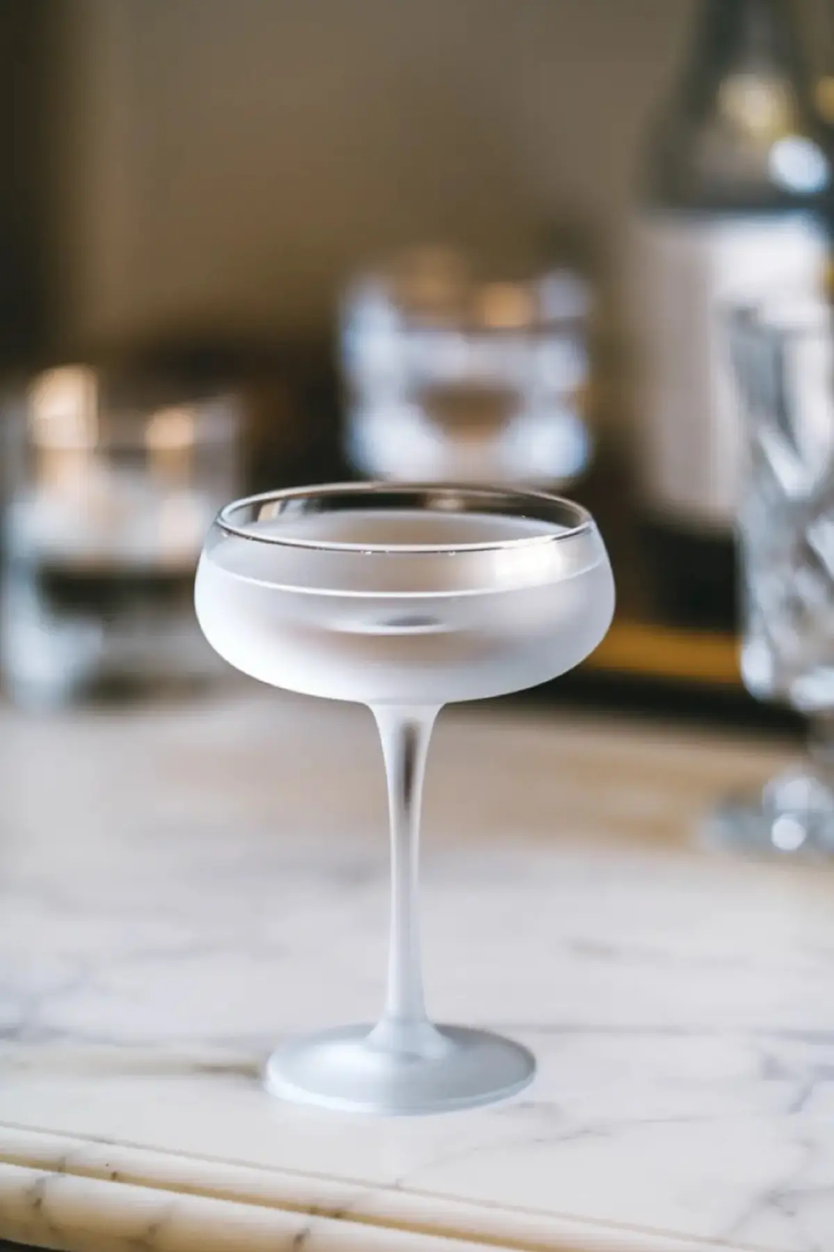 Frosted coupe glass with an elegant, minimalist design, set against a blurred background of barware. The sleek, chilled glass offers a sophisticated, modern look, perfect for serving classic cocktails.